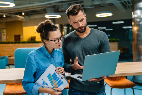 Two helpful young colleagues discussing and using a laptop and diagrams in a modern office, collaborating on a project and sharing ideas in a productive workspace.