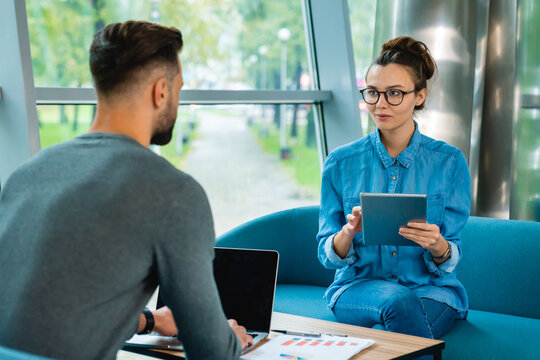 Two young Caucasian colleagues talking together and using gadgets in a cozy office, collaborating and sharing ideas in a comfortable and modern work environment.