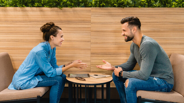 Two colleagues in their 30s sitting and drinking coffee together in the office cafeteria, enjoying a break and engaging in casual conversation in a relaxed work environment.