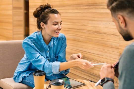 Two young colleagues discussing work while drinking coffee on their break, using a tablet to review tasks and collaborate in a modern office setting.