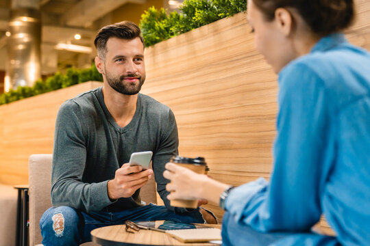 Two young colleagues drinking coffee together during their work break in the office cafeteria, relaxing and chatting in a casual, social work environment.