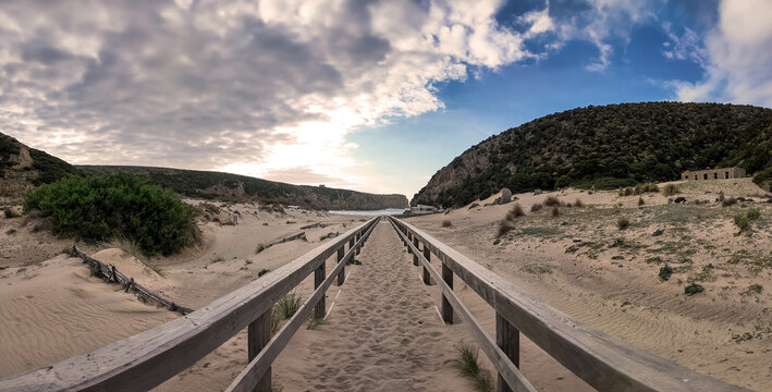 View of Spiaggia di Cala Domestica with a wooden walkway leading through sand dunes and green hills under a cloudy sky Provincia del Sulcis Iglesiente, Sardegna, Italy.