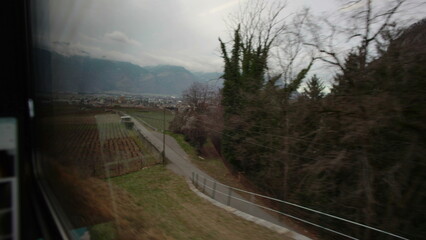 Fototapeta premium Wide valley with vineyards and distant town seen from train window, mountain range rising beyond farmland during countryside rail journey