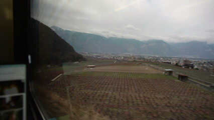 Fototapeta premium Wide valley with vineyards and distant town seen from train window, mountain range rising beyond farmland during countryside rail journey