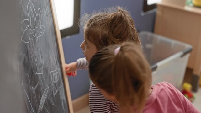 Two young girls draw with chalk on an easel chalkboard and point finger at drawings near a toy storage bin in building; curiosity learning.
