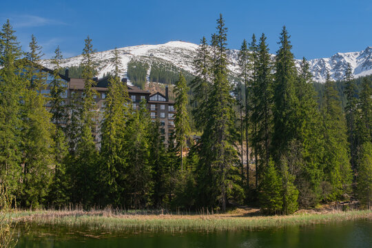 View of Vrbicke pleso lake with dense evergreen forest and a modern hotel with snow-capped Low Tatras mountains in the background Demanovska Dolina, Zilina Region, Slovakia.
