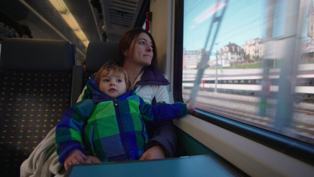 Mother holding toddler beside train window during rail journey, child looking toward camera while parent watches passing countryside in quiet travel moment