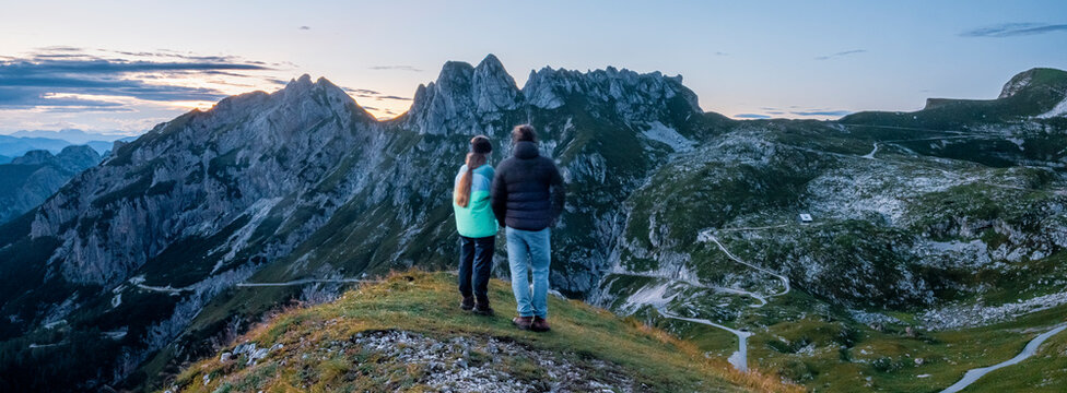 Couple of hikers, man and woman,admiring panoramic mountain peaks at sunset, experiencing outdoor lifestyle and travel adventure on the scenic landscape of Mangart, Julian Alps, Italy-Slovenia border.