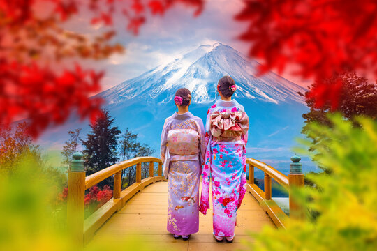 Japanese tourism and cultural travel. Women in kimono stand on wooden bridge viewing Mount Fuji through red autumn leaves.