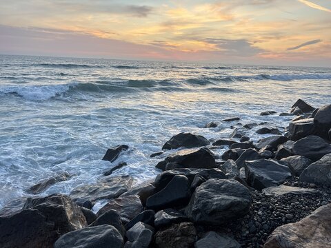 View of ocean waves crashing against dark rocks on the shoreline under a sunset sky in Oceanside, California, United States.