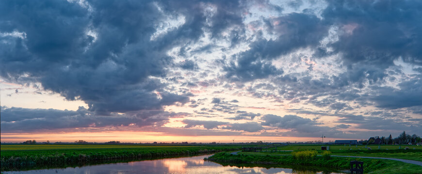 View of polder Spanbroek with a calm canal reflecting a dramatic sunset sky over green fields Spanbroek, North Holland, Netherlands.