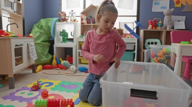 Girl kneeling on foam mat, reaching into a clear plastic bin to sort red and green blocks in a building; concentration learning.