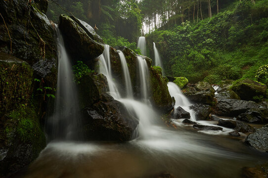View of Air Terjun Grenjengan Kembar cascading over dark mossy rocks surrounded by lush green forest in Kecamatan Pakis, Jawa Tengah, Indonesia.