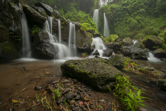 View of Air Terjun Grenjengan Kembar waterfall cascading over dark rocks surrounded by lush green forest in Kecamatan Pakis, Jawa Tengah, Indonesia.