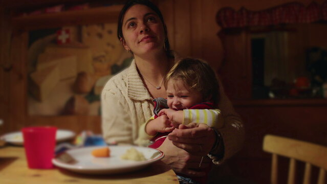 Mother trying to feed toddler at wooden dining table inside cozy alpine chalet, child turning head away and refusing food during intimate family dinner