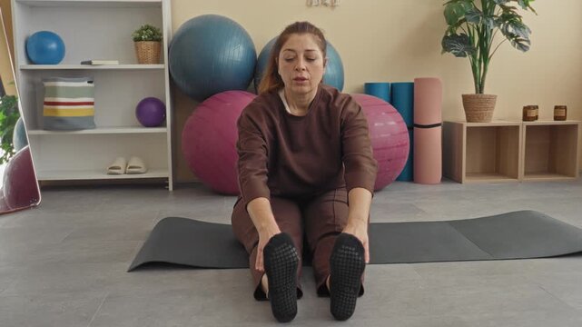 Woman on yoga mat reaches hands to toes while seated, stretching hamstrings in a fitness studio with exercise balls and foam rollers visible; calm focus.