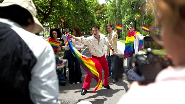 A group of LGBTQ friends danced with rainbow flags during a Pride Month parade on a sunny, tree-lined street, while reporters photographed and reported on the event