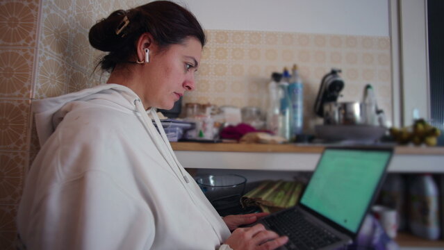 Close side view of woman using laptop at kitchen shelf workspace, concentrated remote worker managing online projects from home environment