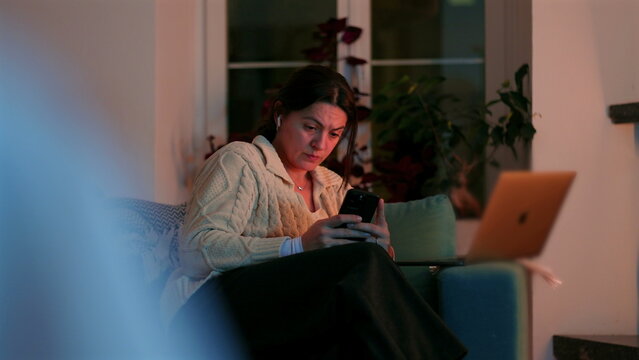Woman sitting on couch at home checking smartphone beside open laptop, managing remote work messages with serious and slightly worried expression in evening