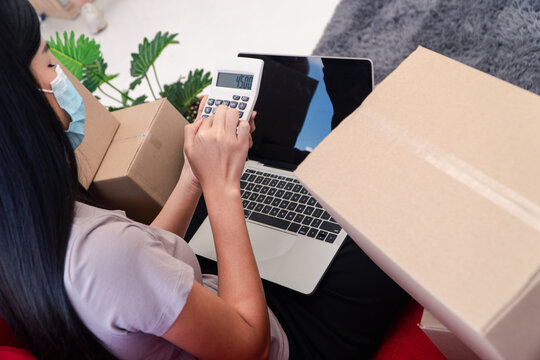 E-commerce Calculations and Delivery: A diligent woman, masked for safety, meticulously calculates shipping expenses, surrounded by cardboard boxes, while a laptop rests nearby.