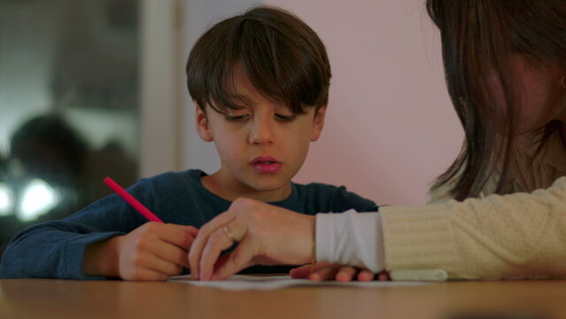 Frustrated mother assisting young son with homework at table correcting mistake and guiding learning during challenging homeschooling session at home