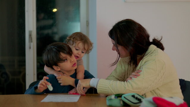 Mother assisting young son with evening homework at table while toddler leans on him capturing genuine candid family moment of schooling and shared learning