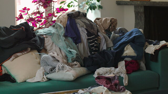 Heaped garments layered across sofa near window and indoor plants portraying chaotic family environment and constant demand of cleaning and organization