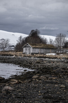 View of a traditional wooden boathouse with a sod roof on a rocky shoreline with snow-capped mountains in the background in Tromso, Troms, Norway.