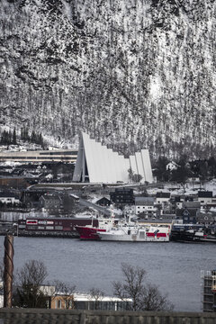 View of the Arctic Cathedral and harbor with ships against a snow-covered mountain backdrop in Tromso, Troms, Norway.