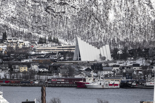 View of the Arctic Cathedral with its iconic triangular architecture against a snowy mountain backdrop and the harbor in Tromso, Troms, Norway.