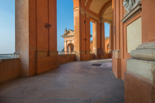 View of the portico and arches of the Santuario Madonna di San Luca with warm terracotta walls and stone flooring under a clear blue sky Bologna, Emilia-Romagna, Italy.