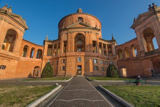 View of Santuario Madonna di San Luca historic circular basilica with its distinctive orange facade, porticoes and Christmas trees in the courtyard, Bologna, Emilia-Romagna, Italy.