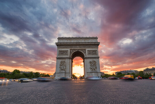 View of the Arc de Triomphe under a dramatic sunset sky with purple clouds and motion-blurred traffic on the cobblestone street in Paris, Ile-de-France, France.