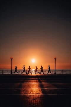 Silhouettes of a group running along a promenade at sunset, with warm light casting long shadows.