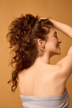 Woman touching curly brunette hair from back on warm orange beauty background. Concept of curl definition, hair volume, natural care, texture treatment and salon styling campaign.