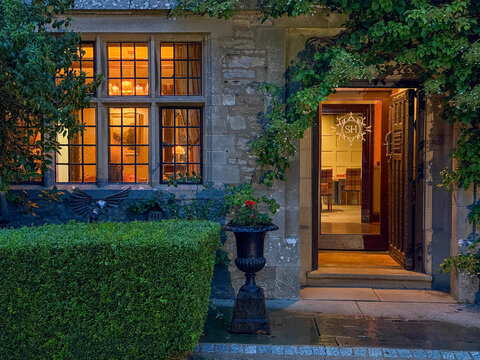 View of a historic stone manor house entrance with warm interior lighting through leaded windows, an open doorway, and lush garden greenery Hawes, England, United Kingdom.