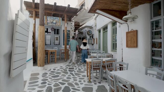 Experience the beauty of Naxos Island in Greece as a couple of men and women stroll through quaint streets lined with traditional architecture. Enjoy the vibrant atmosphere and warm sunshine of summer