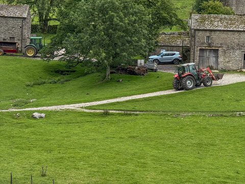 View of a red tractor driving on a gravel path past traditional stone farm buildings and green pastures in Hawes, England, United Kingdom.