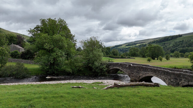 View of an ancient stone arch bridge crossing a flowing river surrounded by lush green meadows and rolling hills under a cloudy sky in Hawes, England, United Kingdom.