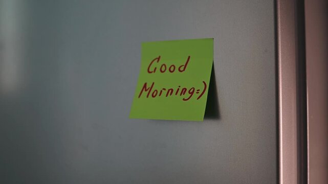 A hand is shown placing a green sticky note on a door with a friendly message. The action takes place indoors during morning time, allowing for a warm start to the day