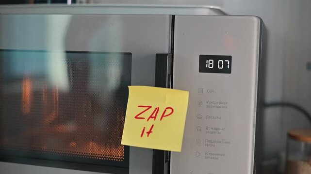 A hand places a sticky note with the word 'ZAP' on a microwave to indicate cooking instructions. The video shows a person giving a thumbs up, signaling approval after using the microwave
