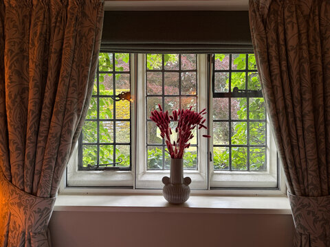 View of a leaded glass window with a vase of red flowers on the sill and patterned curtains looking out onto a lush green garden in Hawes, England, United Kingdom.