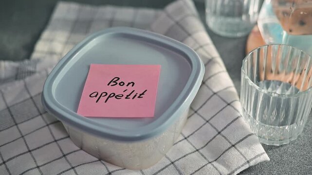 A hand places a note bon appetit on a sealed container with food while glassware sits near by. The setting suggests a meal is being prepared, ready for sharing or serving