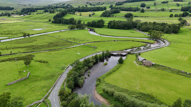View of the winding River Ure and a stone bridge crossing a road amidst the lush green pastures and stone walls of the Yorkshire Dales in Hawes, England, United Kingdom.