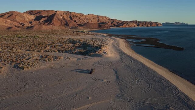 Aerial view shows teardrop camper trailer at sunrise camping on beach near Papa Fernandez in Baja California Mexico with ocean waves and hills nearby
