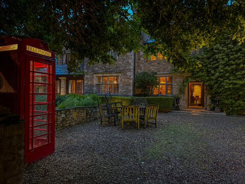 View of an illuminated red telephone booth and a historic stone building with glowing windows and outdoor seating in a gravel courtyard Hawes, England, United Kingdom.
