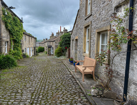 View of a narrow cobblestone street lined with historic stone cottages and a wooden bench under an overcast sky in the Yorkshire Dales Hawes, England, United Kingdom.