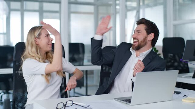 Two Caucasian business colleagues finish complete laptop task dancing giving high five successful teamwork effective cooperation celebrating at office table businesswoman with businessman happy dance