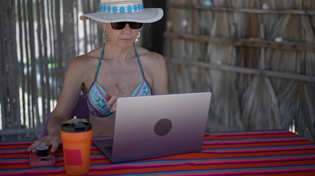 Mature woman shows different emotions as she works on a computer and phone on a tropical beach while relaxing on vacation.