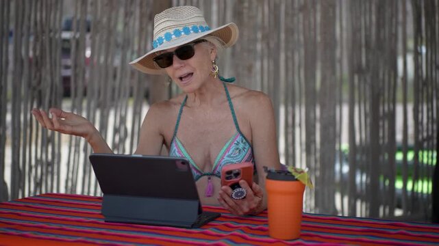 Mature woman has happy video chat on computer tablet, while on a beach during vacation time in a tropical setting.